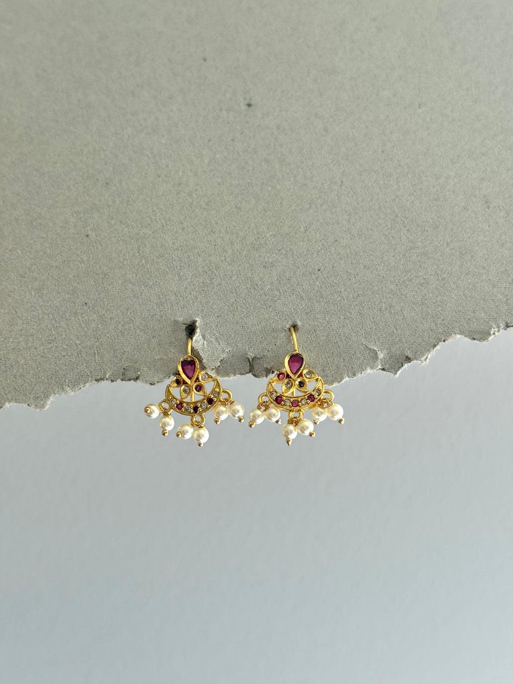 Pair of gold earrings with red gemstones and pearl-like elements on a gray background