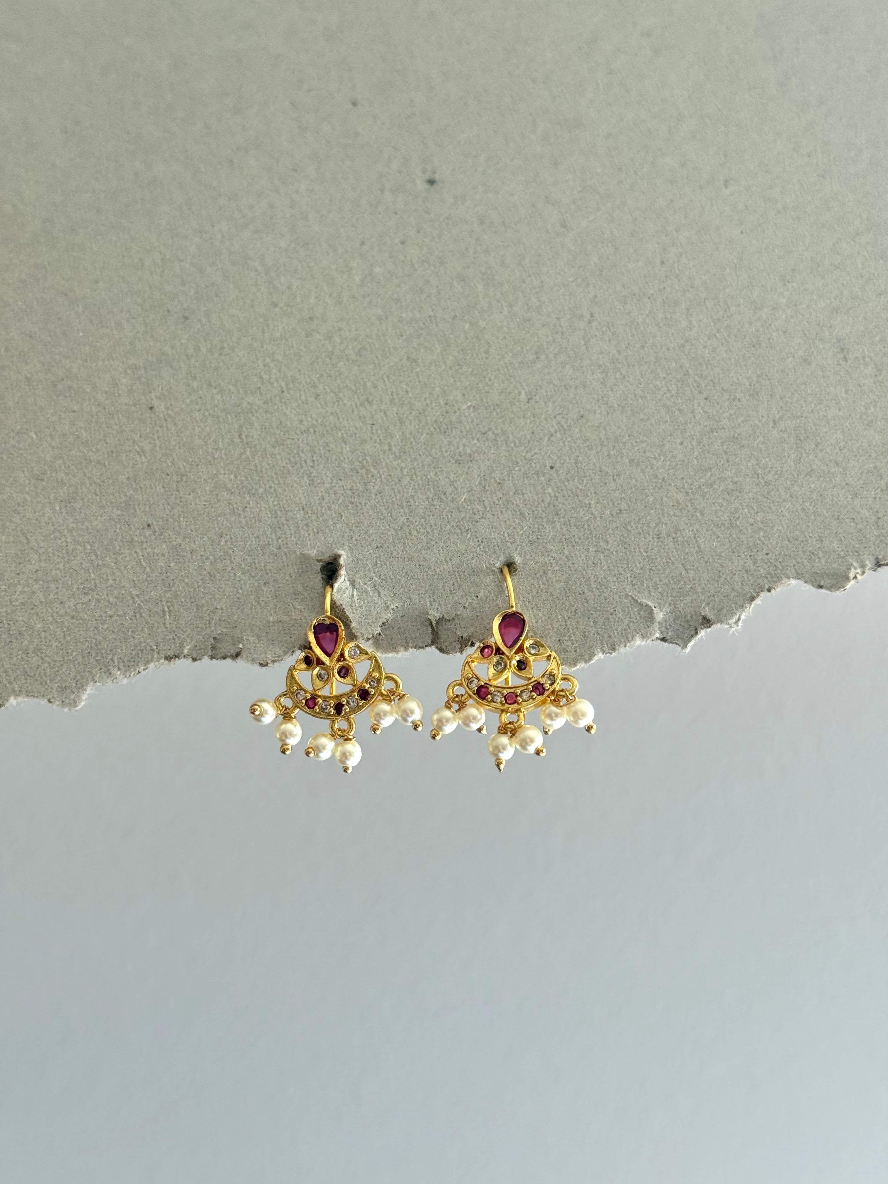 Pair of gold earrings with red gemstones and pearl-like elements on a gray background